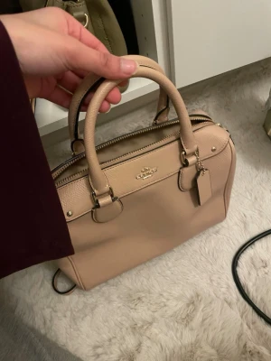 Beige handväska från Coach  - Snygg beige handväska från Coach i  med klassisk form och guldfärgade detaljer. Väskan har två handtag, justerbar axelrem och dragkedja upptill. Perfekt storlek för det viktigaste och stilren look med Coach-logga framtill. Priset går att diskuteras!💕💕
