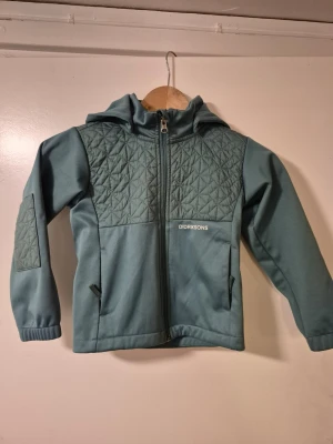 Grön vindjacka Didriksons 110 - Snygg grön vindjacka från Didriksons i storlek 110. Jackan har quiltade partier på bröstet och ärmarna, huva och två fickor med dragkedja. Perfekt för blåsiga dagar och har en stilren look med diskret logga framtill.