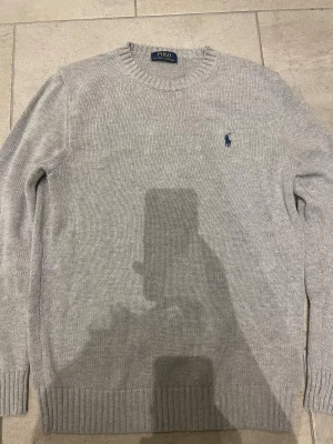 Grå stickad tröja från Polo Ralph Lauren - Snygg grå stickad tröja från Polo Ralph Lauren i 100% bomull. Klassisk rund halsringning, ribbade muddar och diskret blå logga broderad på bröstet. Perfekt för lager-på-lager och enkel att matcha med jeans eller chinos. Tvyörr har storleks lappen åkt bort in tvätten men den va stog S