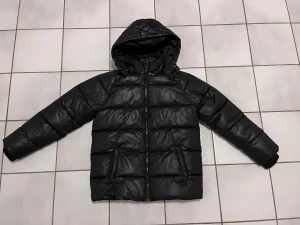 Svart pufferjacka med huva strl 158 - Säljer en svart pufferjacka med huva och dragkedja framtill. Jackan har en quiltad design med breda kanaler och två sidofickor. Perfekt för kalla dagar och har en modern, clean look som funkar till de flesta outfits. Pojk jacka i strl 158!