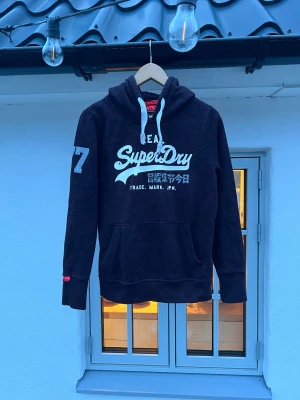 Mörkröd Superdry hoodie med tryck - Snygg mörklila hoodie från Superdry med stort vitt tryck på bröstet och siffran 77 på ena ärmen. Klassisk känguruficka framtill och vita dragsnören i huvan. Mjuk bomullsblandning som är skön att ha på sig. Perfekt för dig som gillar streetwear och sportig stil.
