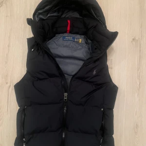 Svart dunväst från Polo Ralph Lauren - Svart dunväst från Polo Ralph Lauren med huva och dragkedja framtill. Klassisk design med diskret logga på bröstet och två fickor med dragkedja. Perfekt för lager-på-lager och snygg till både hoodie och t-shirt. Skulle säga att det är perfekt nu när det börjar bli kallare. Skriv vid minsta fundering!!
