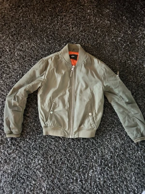 Olivgrön bomberjacka med dragkedja - Snygg olivgrön bomberjacka med klassisk orange foder. Jackan har ribbade muddar, två fickor med tryckknappar och en dragkedja framtill. Perfekt för dig som gillar streetstyle och vill ha en jacka med cool attityd.