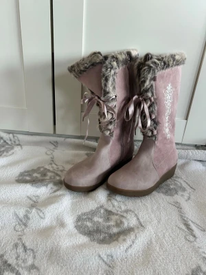 Rosa mockastövlar med päls och broderi - Säljer ett par ljusrosa stövlar med mjuk mocka och fluffig grå päls runt skaftet. Stövlarna har dekorativt vitt broderi på utsidan och snygga satinband som snörning. Sulan är platt och brun, perfekt för vinter och höst. Unik och mysig look!