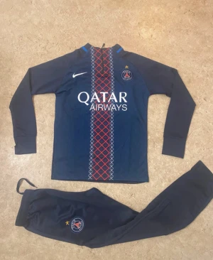 PSG träningsset Nike marinblå - Säljer ett PSG träningsset från Nike i marinblått med långärmad tröja och matchande byxor. Tröjan har PSG-logga, Nike swoosh och Qatar Airways-tryck samt rött och vitt mönster framtill. Byxorna har PSG-emblem och resår i midjan. Perfekt för fotbollsträning.