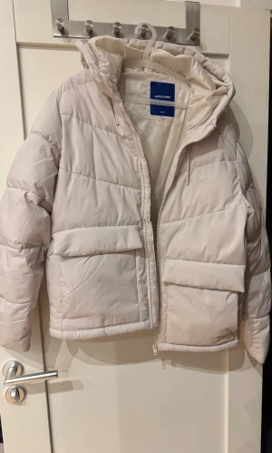 Vit pufferjacka från Jack & Jones - Säljer en snygg vit pufferjacka från Jack & Jones med huva och stora fickor framtill. Jackan har quiltad design och dragkedja hela vägen upp. Perfekt för kalla dagar och har en clean look som passar till streetwear.