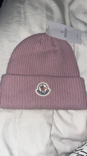 Ljusrosa Moncler mössa i ull - Snygg ljusrosa ribbstickad mössa från Moncler med klassisk logga framtill. Mössan har uppvikt kant och är tillverkad i mjuk ull som håller dig varm. Perfekt accessoar för att lyfta din vinterstil.