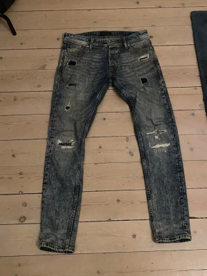 Blå slitna skinny jeans med hål - Säljer ett par blå skinny jeans med coolt slitna detaljer och flera hål på framsidan. Jeansen har en tvättad look och klassisk femficksdesign.  Dom är storlek 29-32 bra skick förutom en lite defekt ( tredje bilden) två hål har blivit ett men går lätt å fixa på några sekunder. Märke-Jack Jones   pris-149