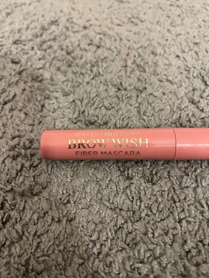 Makeup Mekka Brow Wish Fiber Mascara - Säljer en Brow Wish Fiber Mascara från Makeup Mekka. Förpackningen är i en snygg matt rosa färg och har en böjd borste för enkel applicering. Perfekt för att ge fylliga och definierade bryn med fiberformula. Tuben är i plast och har guldfärgad text.