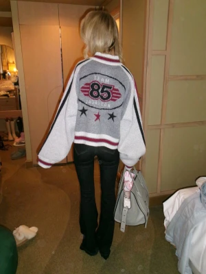 Grå kofta med dragkedja  - Säljer en oversized grå kofta från Jaded London med dragkedja framtill och coolt Team 85-tryck på ryggen. Tröjan har breda ärmar, röd och svart rand längs ärmarna och ribbade muddar i vitt, rött och svart. Perfekt för dig som gillar streetstyle och sportig vibe.