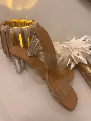 Klara sandaler med transparent klack - Säljer ett par trendiga sandaler med beige sula och breda, transparenta remmar. Skorna har fyrkantig tå och hög, klar akrylklack som ger en modern vibe. Perfekta för dig som vill sticka ut med en clean och stilren look.