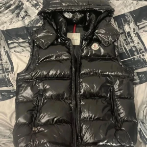 Svart dunväst från Moncler - Snygg svart dunväst från Moncler med glansig finish och quiltad design. Västen har huva, två fickor med dragkedja och klassisk Moncler-logga på bröstet. Perfekt för lager-på-lager och streetstyle. Dragkedja framtill och coolt märke på insidan.