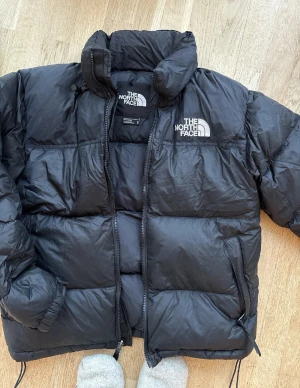 1996 RETRO NUPTSE JACKET UNISEX - Dunjacka  - 1996 RETRO NUPTSE JACKET UNISEX - Dunjacka - black Säljer en svart dunjacka från The North Face med dragkedja och broderad logga på bröstet. Jackan har en hög krage och är perfekt för kalla dagar. Modellen är klassisk och tidlös, vilket gör den till ett måste i garderoben. 