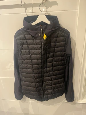 Svart pufferjacka från Parajumpers - Snygg svart pufferjacka från Parajumpers med huva och quiltad framsida. Jackan har dragkedja, säkerhetsficka med orange detalj och stilrena svarta paneler på ärmarna. Perfekt för dig som vill ha en cool och funktionell jacka med streetkänsla.