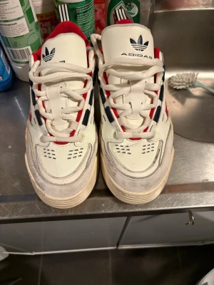 Adidas Continental 80 sneakers vit/röd - Säljer ett par Adidas Continental 80 sneakers i vitt med röda och marinblå detaljer. Skorna har klassiska ränder på sidan, snörning och en platt sula med mönstrad undersida. Insidan är fodrad med rött tyg och ovandelen är i skinn och syntet. Perfekt för dig som gillar retrostil.