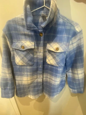 Blåvit rutig overshirt från Lindex - Säljer en blå och vit rutig overshirt från Lindex med två bröstfickor och snygga knappar framtill. Jackan har en klassisk krage och långärmade ärmar, perfekt för lager-på-lager. Materialet är mjukt och känns skönt mot huden.