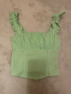 Grön volangtop från Gina Tricot XS - Supersöt ljusgrön topp från Gina Tricot i storlek XS. Toppen har breda axelband med volangdetaljer och är tillverkad i ett ribbat, lätt skrynkligt syntetmaterial. Croppad passform som ger en fräsch och trendig look, perfekt för vår och sommar.