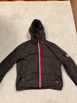 Svart vindjacka från Moncler - Svart vindjacka från Moncler med huva och dragkedja framtill i rött, vitt och blått. Jackan har två fickor med dragkedja och Moncler-logga på ärmen. Tillverkad i lätt polyester, perfekt för dig som vill ha en clean och sportig look.