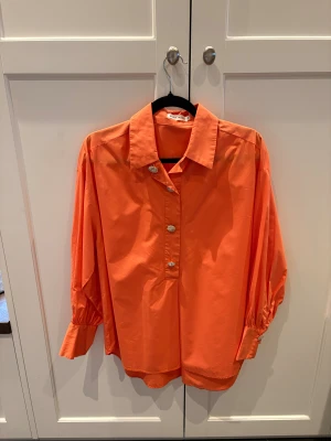 Orange skjorta med glittriga knappar - Snygg orange skjorta med glittriga dekorationsknappar framtill. Skjortan har en klassisk krage, långa ärmar med breda manschetter och en loose passform. Perfekt för dig som vill sticka ut med färg och coola detaljer. Inköpt på Two Angela i Göteborg 
