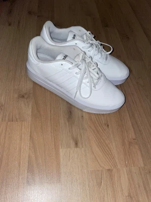 Vita sneakers från Adidas - Fräscha vita sneakers från Adidas med klassiska tre ränder på sidan och snörning framtill. Skorna har en rund tå och platt sula, tillverkade i syntetmaterial med insida i textil. På innersulan står det 'End Plastic Waste', vilket visar att de är miljövänliga.