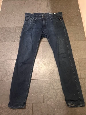 Replay Anbass mörkblå jeans slim fit - Snygga mörkblå Replay Anbass jeans med slim fit och klassisk femficksdesign. Jeansen har raka ben, normal passform och är tillverkade i mjukt jeansmaterial med diskreta slitningar. Perfekta för dig som gillar stilrena och tidlösa jeans. 34 W 32 L