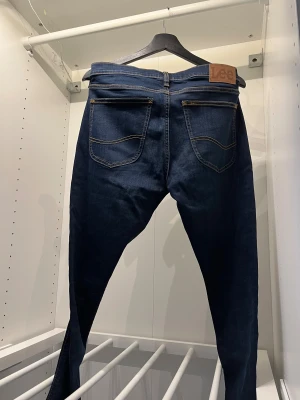 Mörkblå slim  jeans från Lee - Snygga mörkblå jeans från Lee med klassisk femficksdesign och kontrastsömmar. Modellen är skinny med smal passform och har en läderpatch med Lee-logga bak i midjan. Jeansen är tillverkade i stretchigt denim för extra komfort. W31 L32