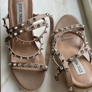 Steve Madden sandaler med nitar - Beige sandaler från Steve Madden med höga flätade platåklackar och flera remmar dekorerade med silvriga nitar. Snyggt edgy look med öppen tå och justerbar rem runt ankeln. Perfekt för dig som gillar statement-detaljer och vill sticka ut.