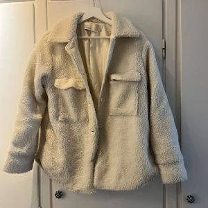 Vit teddyjacka från NLY Trend - Vit/cream teddyjacka från NLY Trend. Jackan har stora bröstfickor med lock, knäppning framtill med knappar och fickor på sidan. 