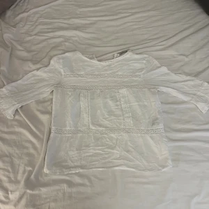 Vit blus med spets från - Säljer en söt vit blus från ONLY i storlek 34. Blusen har trekvartsärm med lätt rynk vid ärmslut, spetsdetaljer och broderade partier framtill. Materialet är mjukt och luftigt, perfekt för vår och sommar. Rund halsringning med spetskant ger en romantisk vibe.