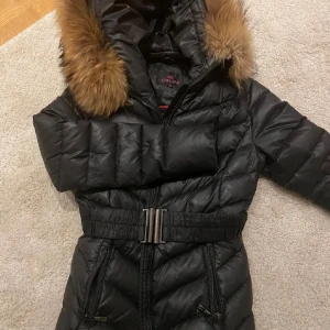 Svart pufferjacka med päls från Chelsea - Säljer en svart pufferjacka från Chelsea med snyggt bälte i midjan och dragkedja framtill. Jackan har huva med brun päls och två dragkedjefickor. Perfekt för kalla dagar och riktigt skön att ha på sig.