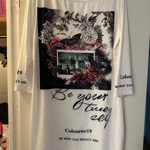 Vit tunika strl: L - Vit tunika med unikt tryck framtill som visar blommor, fjärilar och stadsmotiv. Texten 'Be your true self' och 'Be who you really are' finns tryckt på både framsidan och ärmarna. Tunikan har lång ärm och är i mjuk bomull.