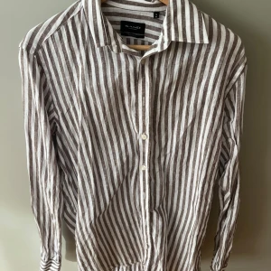 Randig linneskjorta från Sand Copenhagen - Snygg randig skjorta från Sand Copenhagen i vitt och brunt. Skjortan är tillverkad i luftig linne, har klassisk krage, långa ärmar och knäppning framtill. Perfekt för en stilren och avslappnad look.