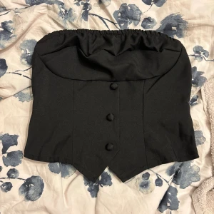 Svart korsett-topp med knappar - Snygg svart korsett-topp med markerad midja, dekorativa knappar framtill och dragkedja bak. Toppen är axelbandslös med elastisk kant upptill för extra passform. Perfekt för att skapa en edgy look.