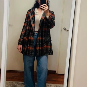 Rutig kappa i orange och svart - Säljer en lång, rutig kappa i orange, svart och beige med stora fickor framtill. Jackan har en rak passform och är tillverkad i ett ull-liknande material. Perfekt för höst och vinter, med snyggt rutmönster som sticker ut.