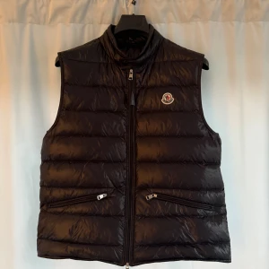 Svart Moncler Gui  - Ett stilrent plagg för dig som är ute för de lilla extra. Denna väst är i storlek 4 motsvarar L och är såklart äkta. Köptes på deras officiella hemsida. Är i ny skick som ni kan se på bilderna. Har bara använts ett få tal gånger. Säljs pågrund av att den inte passar längre. Dustbag,Tags och kvitto finns! Ett rimligt pris för de du får  