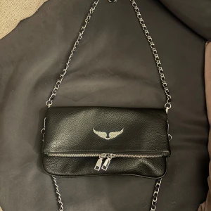 Zadig väska  - Snygg svart axelväska i skinn med silverfärgade kedjedetaljer och dragkedja framtill. Väskan har ett stilrent emblem med vingar i silver på framsidan och magnetlås. Perfekt storlek för det viktigaste och ger en cool vibe till din outfit.