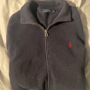 Mörkblå stickad kofta från Polo Ralph Lauren - Snygg mörkblå stickad kofta från Polo Ralph Lauren med dragkedja och klassisk röd broderad logga på bröstet. Koftan har ribbad struktur, ståkrage och praktiska fickor framtill. Perfekt för dig som gillar stilrena och tidlösa plagg. Pris kan diskuteras!!