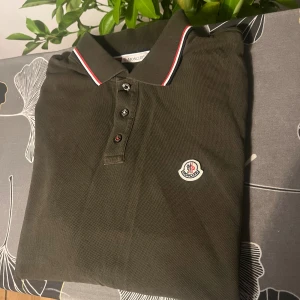 Mörkgrön Moncler pikétröja - Snygg mörkgrön pikétröja från Moncler med klassisk krage och tre knappar framtill. Tröjan har Moncler-logga broderad på bröstet och kragen är dekorerad med röd, vit och marinblå rand. Perfekt för dig som gillar stilrena märkesplagg.  Längd 62cm bredd 48cm