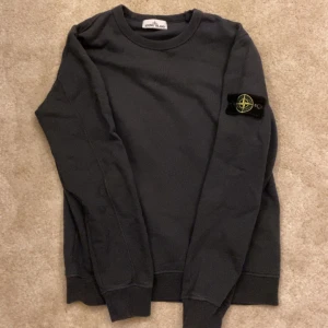 Grå sweatshirt från Stone Island - Säljer en svart långärmad sweatshirt från Stone Island med den klassiska loggan på vänster ärm. Tröjan har rund halsringning och ribbade muddar vid ärmslut och nederkant. Perfekt för dig som gillar streetwear och stilren design. Köpte av en kompis så osäker på äkthet.