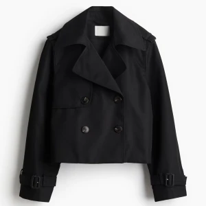 Svart croppad trenchcoat  - Snygg svart croppad trenchcoat med bred krage, dubbelknäppning och bältesdetaljer vid ärmsluten. Jackan har en stilren och modern look som passar perfekt till streetwear och trendiga outfits. Slutsåld på hemsidan prislapparna kvar!