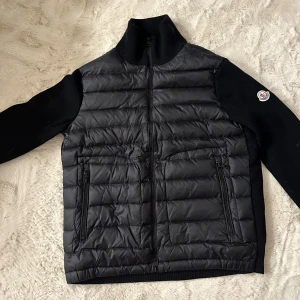 Svart Moncler cardigan - Svart cardigan från Moncler med stickade ärmar och hög krage. Jackan har två dragkedjeförsedda fickor fram och ikonisk Moncler-logga på vänster ärm. Materialmix med quiltad polyester på kroppen och stickad ull på ärmar och krage. Perfekt för kyliga dagar. Passar L inget mindre.