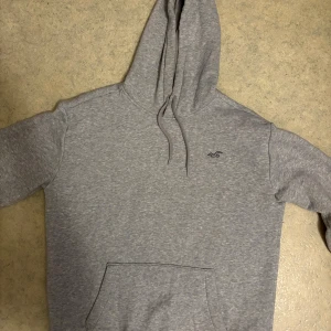 Grå hoodie från Hollister med luva - Säljer en stilren grå hoodie från Hollister med klassisk huva och magficka. Tröjan har dragsko i halsen och diskret Hollister-logga på bröstet. Perfekt för chill dagar och enkel att matcha med jeans eller joggers.