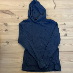 Blå hoodie från Polo Ralph Lauren - Säljer en blå hoodie från Polo Ralph Lauren i ribbad bomull. Tröjan har huva, lång ärm och klassisk broderad logga på bröstet. Perfekt för dig som gillar en avslappnad och stilren look.