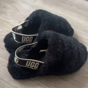 Svarta fluffiga sandaler från UGG - Ugg fluff yeah tofflor. Supermjuka och fluffiga svarta sandaler från UGG med bred resårrem med logga över hälen. Perfekta för dig som vill ha något riktigt cozy och trendigt på fötterna. Endast använt 2 gånger men lite dammiga