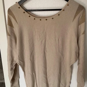 Beige tröja med gulddetaljer - Beige långärmad tröja med guldfärgade detaljer på axlarna och små nitar runt den breda halsringningen. Tröjan är tillverkad i en mjuk blandning av viskos och nylon och har ribbade muddar vid ärmslut och nederkant. Perfekt för dig som gillar stilrena plagg med en twist.