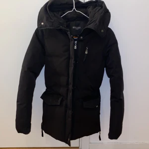 Svart dunjacka från Hollies med huva - Säljer en svart dunjacka från Hollies med stor, mysig huva och dragkedja framtill. Jackan har flera fickor med lock och dragkedja, samt justerbar midja för snygg passform. Perfekt för kalla dagar och riktigt stilren look.