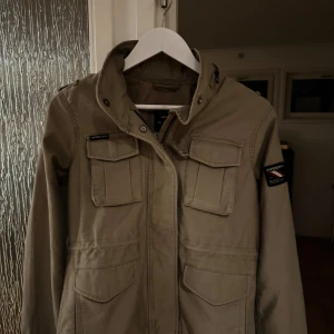 Beige parkajacka från Superdry  - Snygg beige parkajacka från Superdry med fyra stora fickor framtill, hög krage och patch på ärmen. Jackan har en rak passform och är tillverkad i bomullsmaterial. Perfekt för dig som gillar en clean och stilren look. Storleken är XS men sitter som S. Är en Dam jacka!
