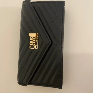 Svart quiltad axelväska Cavalli Class - Snygg svart axelväska från Cavalli Class i quiltat skinn med guldfärgad kedja och logga framtill. Väskan har ett stilrent mönster i v-format och stängs med tryckknapp. Helt ny!