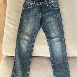 Jack & Jones Slim Glenn jeans blå - Snygga blå jeans från Jack & Jones, modell Slim Glenn. Jeansen har en slim passform, klassisk femficksdesign och knäppning med guldiga knappar. Materialet är jeans med lätt slitning och kontrastsömmar för en modern look.