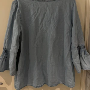 Blå jeansblus med trumpetärm - Säljer en ljusblå blus i jeansmaterial med snygga trumpetärmar och rynkade detaljer vid ärmsluten. Blusen har en enkel knapp i nacken och en rak, avslappnad passform. 